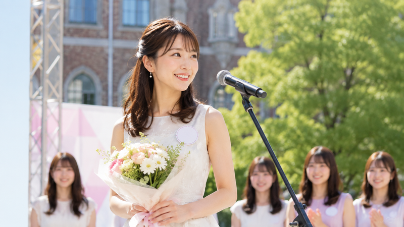 京都の大学キャンパスをイメージした背景で、大学生の女性がステージに立っている様子。華美になりすぎず、清楚で爽やかな雰囲気