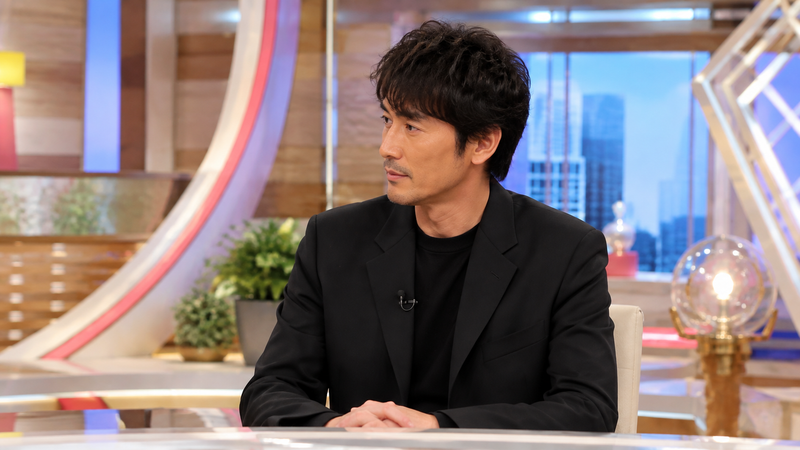 ニュース・情報番組のスタジオを背景に、黒系の衣装を着た長身の男性俳優が出演している場面。