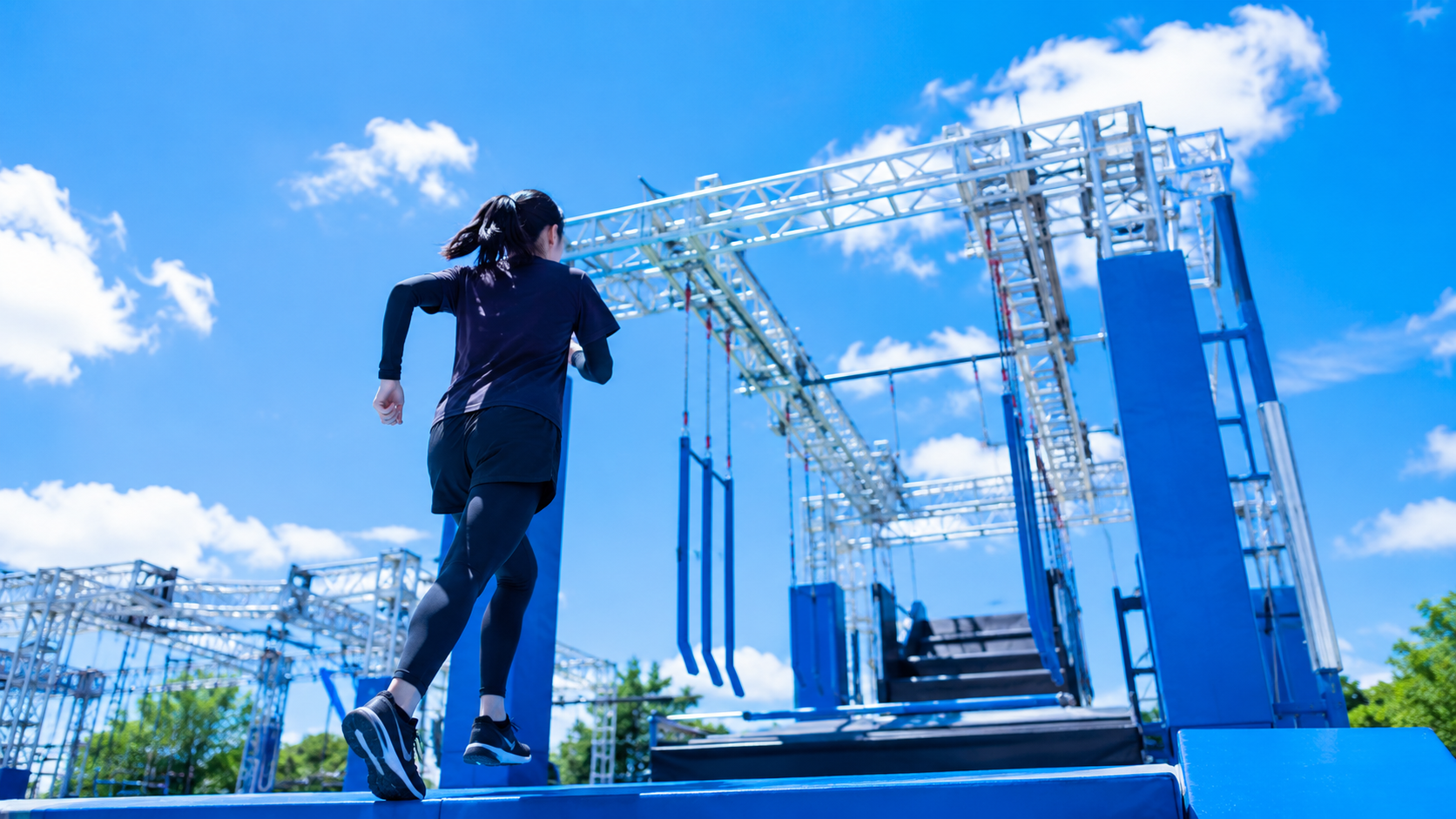 青い大型アスレチックを背景に、障害物へ向かう女性選手のシルエット。爽